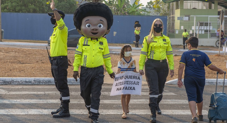 Agentes de trânsito, juntamente com as mascotes ‘Agente Vidinha e Amarelinho’, realizando a travessia de pais e alunos na faixa de pedestre